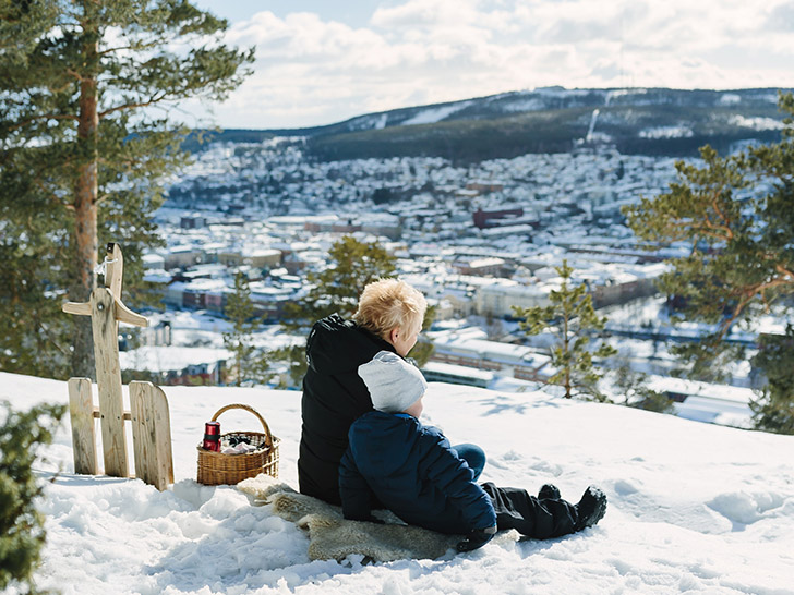 Vuxen med barn sitter på Norra Berget i vintermiljö med utsikt över Sundsvall. Foto: Norra Berget.