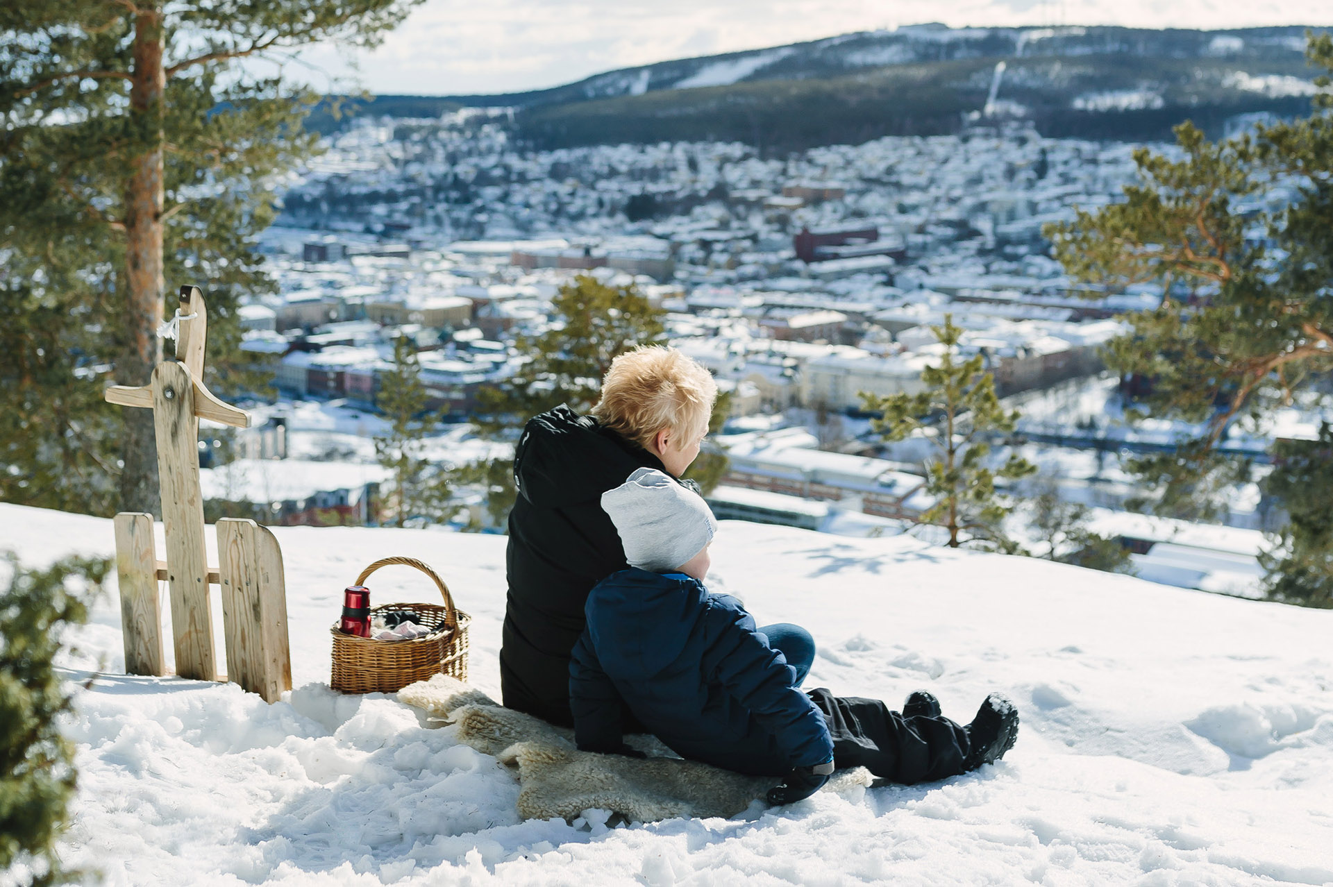 Vuxen med barn sitter på Norra Berget i vintermiljö med utsikt över Sundsvall. Foto: Norra Berget.